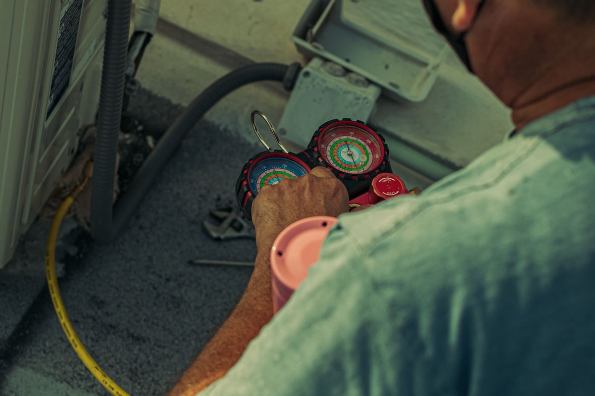 Refrigerant manifold gauges in a technician's hand during an air conditioner repair at an outdoor unit.